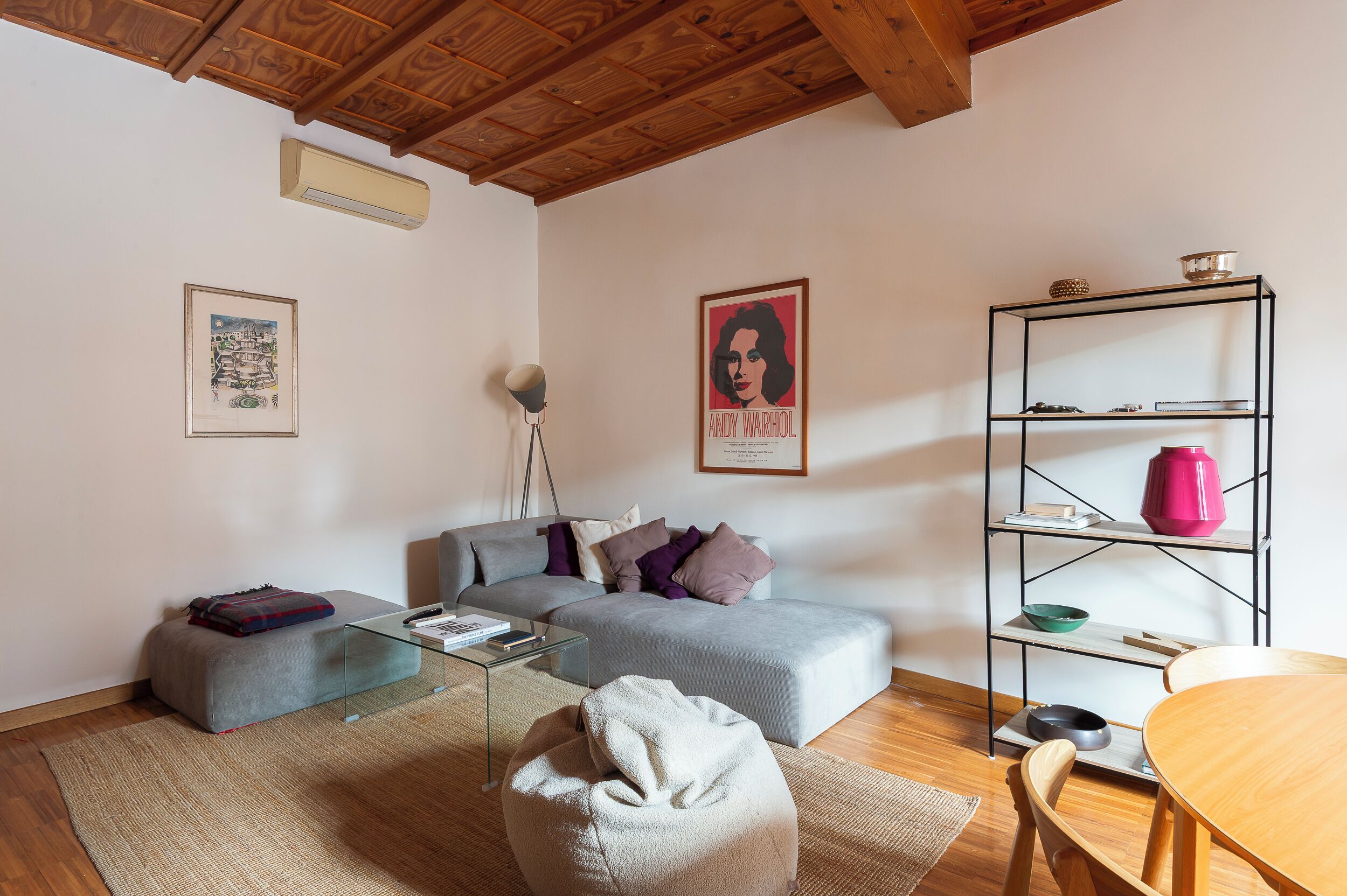 A modern living room with a grey sofa, a glass coffee table, a wooden dining table, and a metal shelving unit with various items.