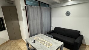 Une pièce avec une table grise, deux chaises, un canapé noir, un rideau gris et une horloge au mur.