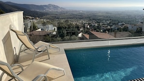 Ein Poolbereich mit Liegestühlen, freie Sicht auf eine Stadt und Berge in der Ferne sowie ein gefliestes Schwimmbecken.