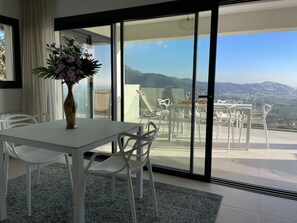 A modern room with a white table and chairs, a vase with flowers, and a balcony with a view of the countryside. - Ref. Ali12 Modern villa with private pool and sea view (Rosas / Roses)