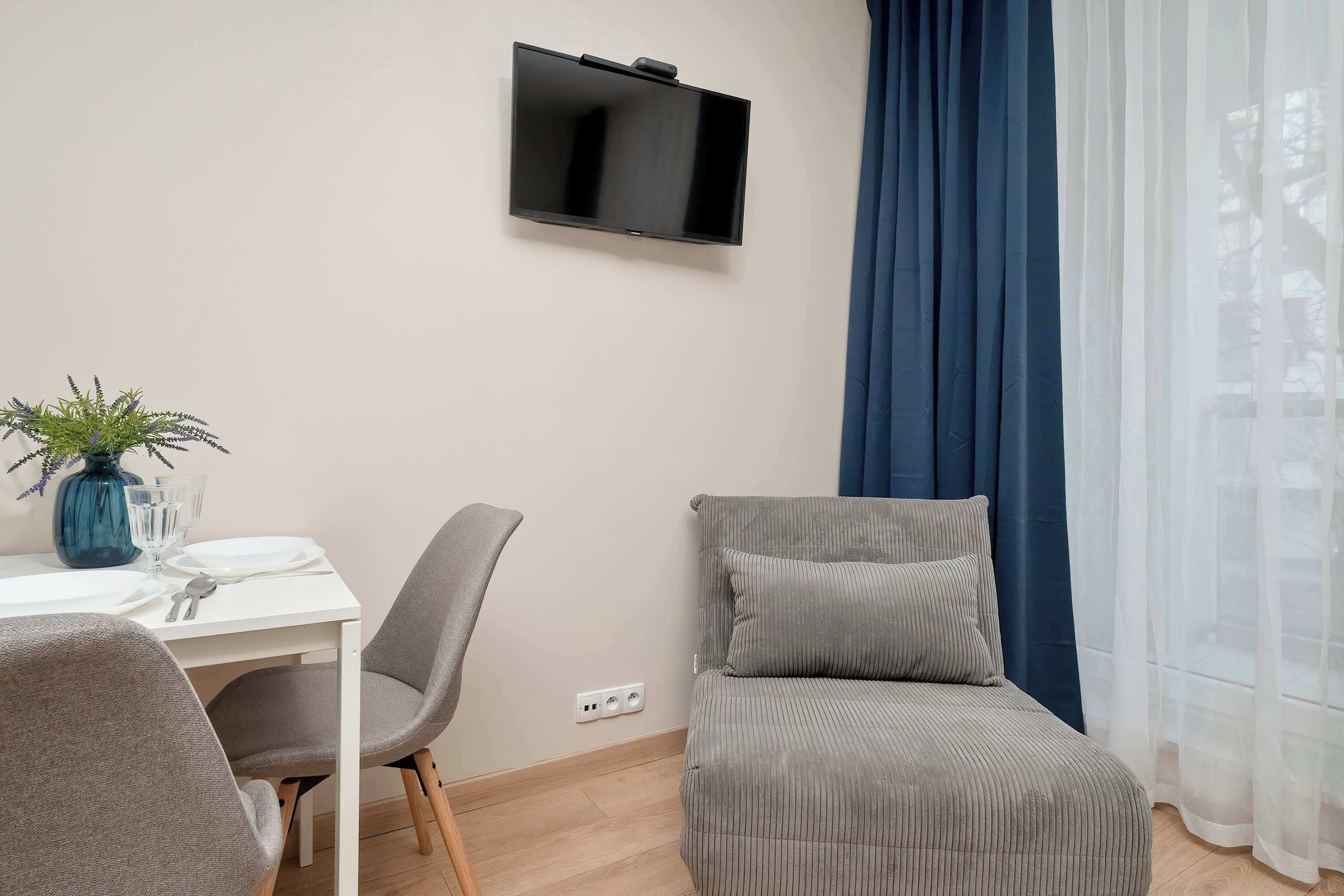 Una habitación con una televisión de pantalla plana en la pared, una mesa de comedor con sillas, una planta pequeña en maceta y un sofá con cojines.
