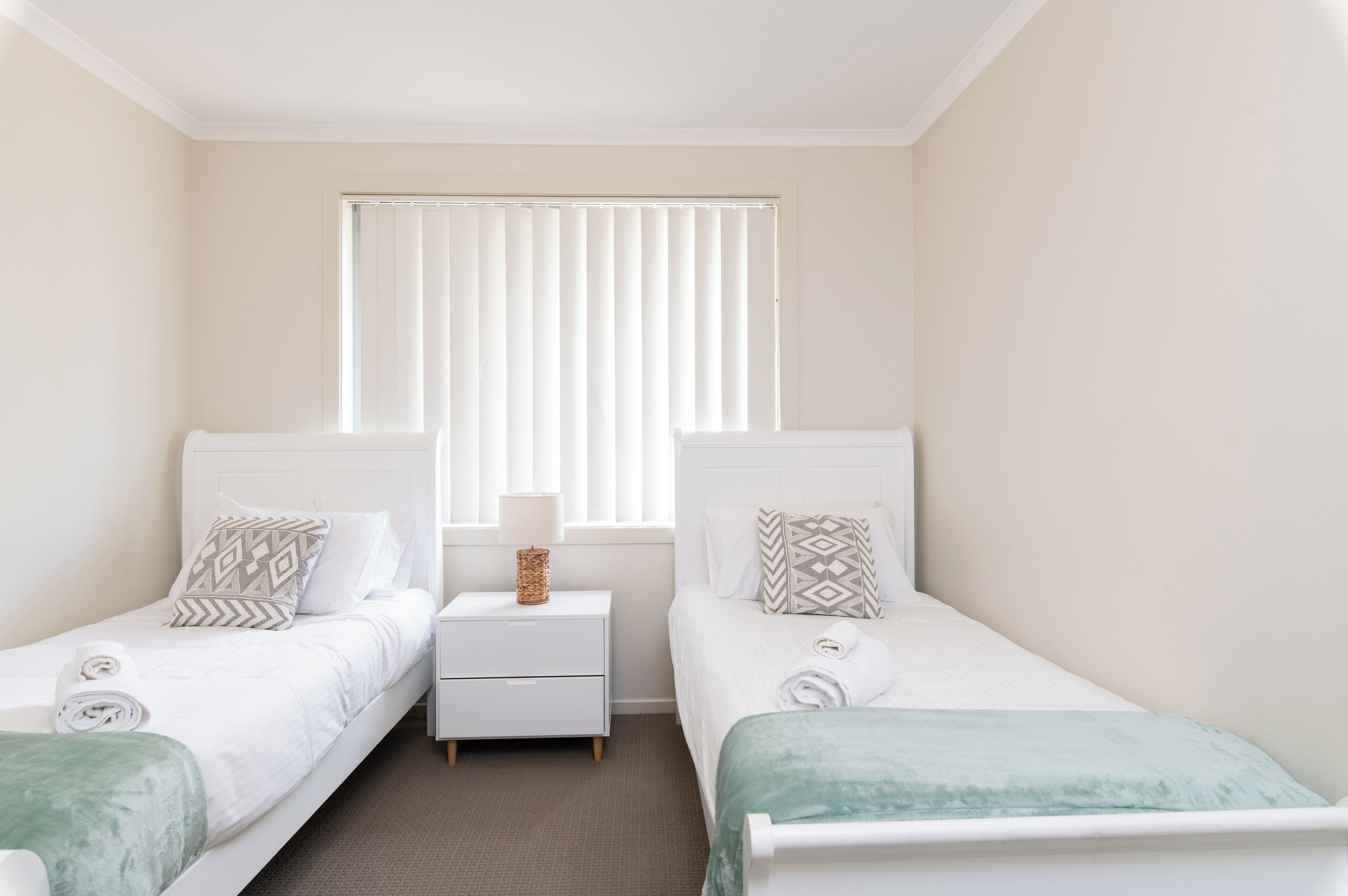 A bedroom with two beds, a nightstand, a window with blinds, and a lamp.