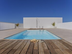 Una piscina rettangolare con un pontile in legno, due piante in vaso e un paio di occhiali da sole e un libro sul pontile.