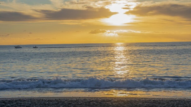 A beach scene with the sun setting over the ocean, boats in the distance, and waves approaching the shore. - CHL El Faro, next to the Puerto Naos beach (Puerto Naos, Los Llanos de Aridane)
