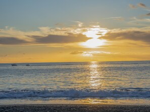 Una spiaggia con il sole che tramonta sull'oceano, barche all'orizzonte e onde che si infrangono sulla riva.