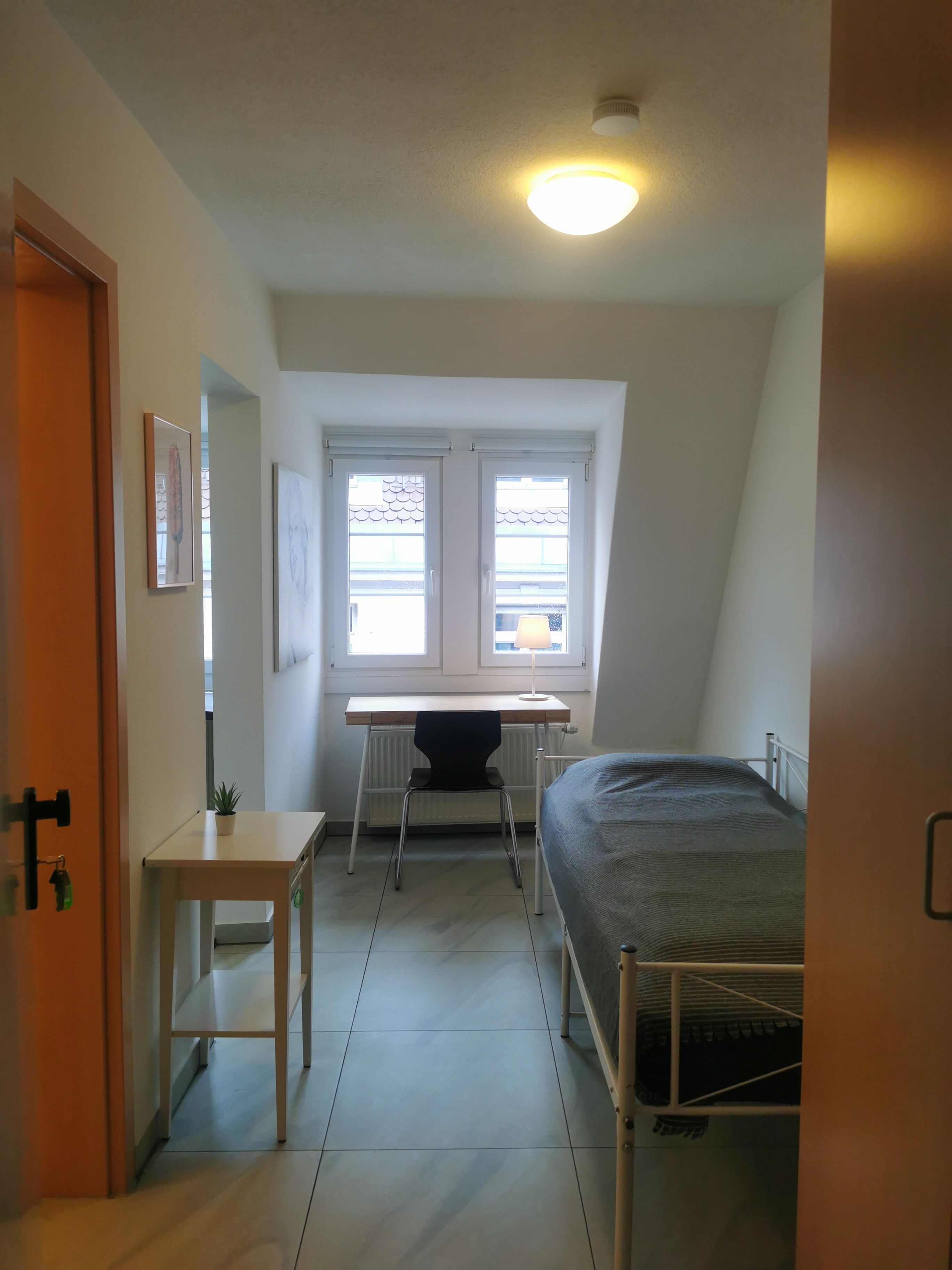 Ein kleiner, gut beleuchteter Raum mit einem Bett, einem Schreibtisch und zwei Fenstern.
