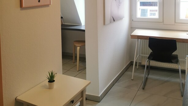 Una habitació amb escriptori, una cadira, una taula petita amb una planta i dos quadres al mur.