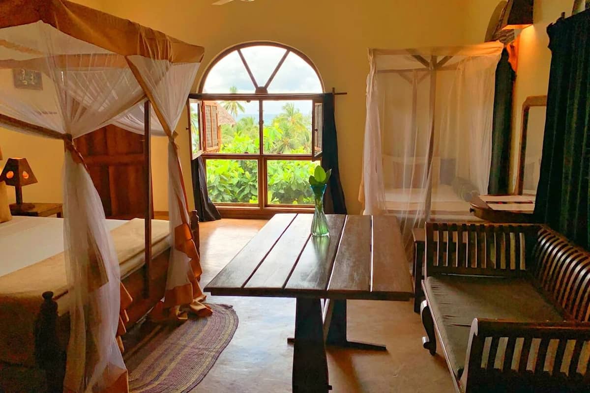 Una habitación con una cama, una mesa de madera, un banco acolchado, un ventanal arqueado grande y una mosquitera.