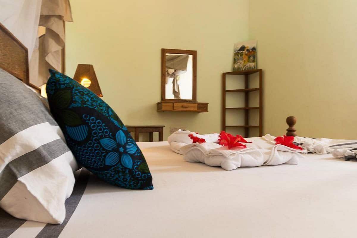 Una cama bien hecha con almohadas, una mesita de noche con una lámpara, un espejo y una estantería con un cuadro.