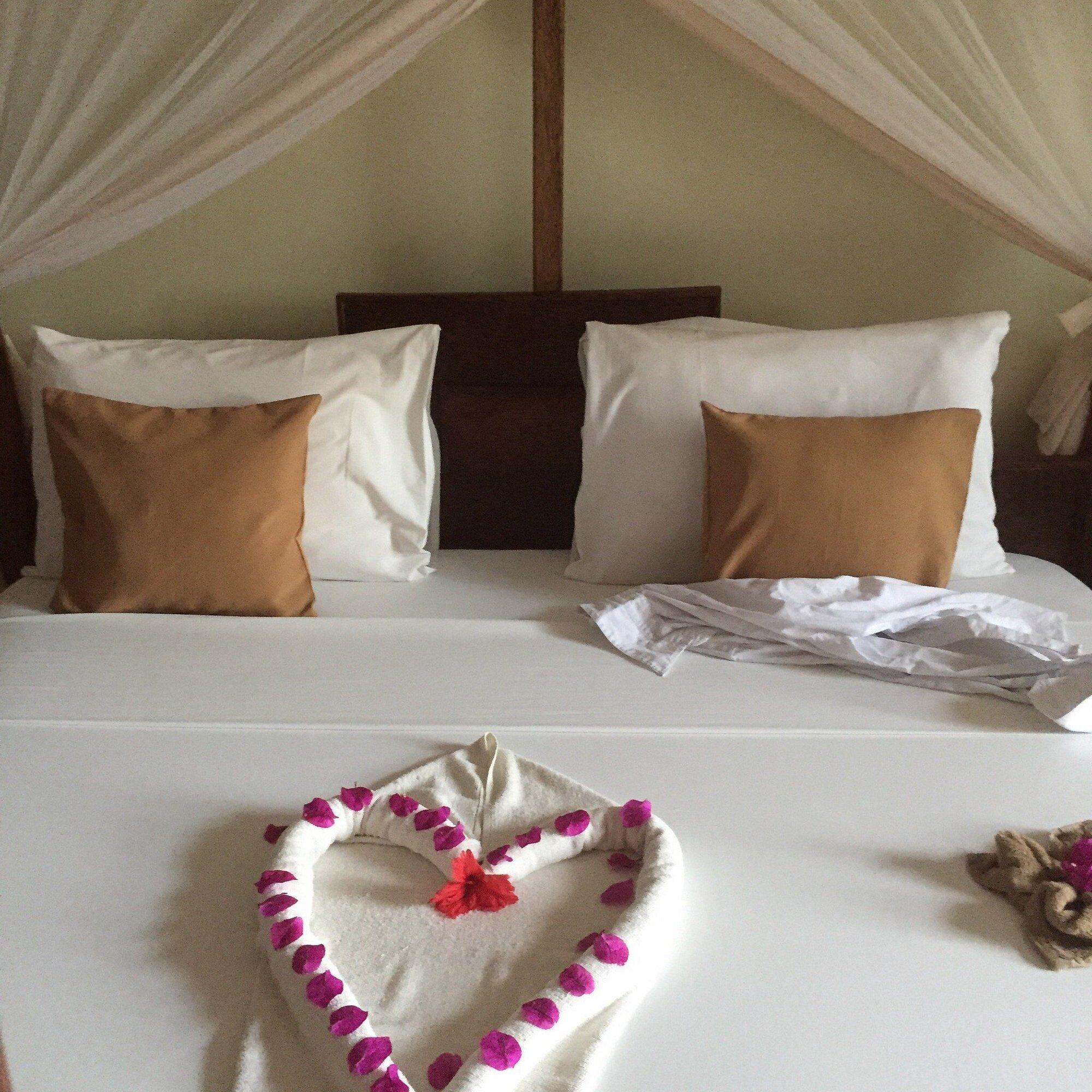 Een bed met witte lakens, twee bruine kussens en een decoratie in de vorm van een hart met roze bloemblaadjes.