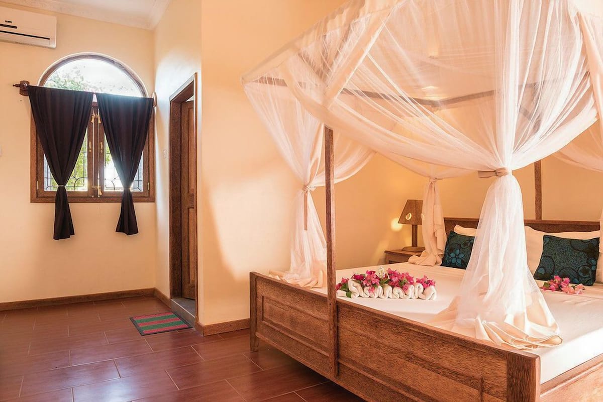 Una cama con dosel y largueros, cabecera de madera, mesita de noche con lámpara y ventana con cortinas.