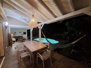 Zona pranzo all'aperto con tavolo e sedie in legno, lampadario e vista su una piscina illuminata di notte.