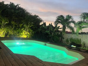 Una piscina rettangolare con un bordo in legno, circondata da una vegetazione tropicale rigogliosa e un recinto in legno.