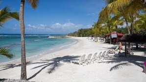 En strand med vit sand, palmer och kristallklart blått vatten. Här finns solstolar och parasoller under ett skuggigt område.