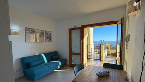 En stue med en blå sofa, et træspisebord med en fjernbetjening, og udsigt til bybilledet gennem en åben dør.