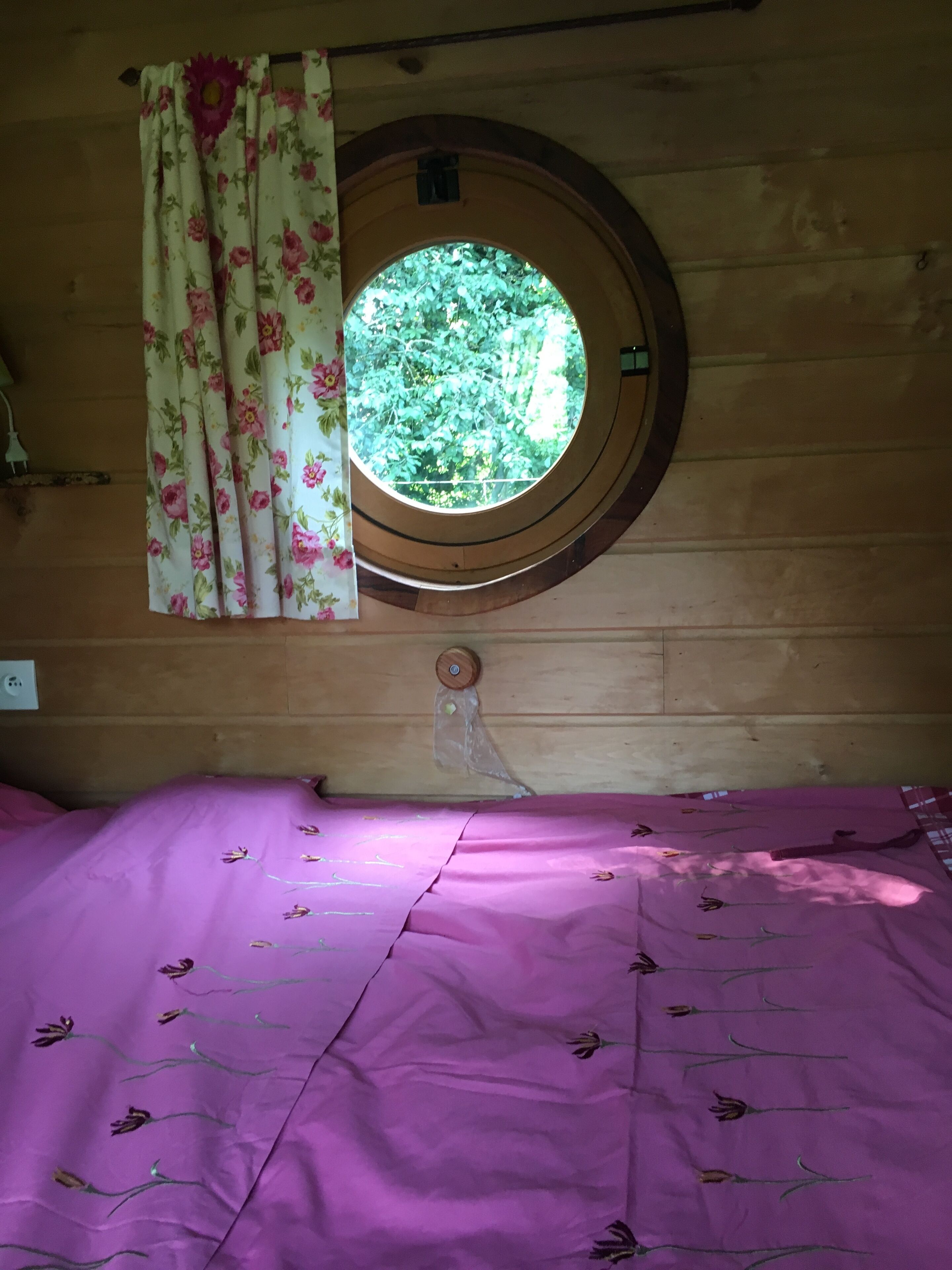 A room with a wooden wall, a round window with a view of greenery, a bed with a purple bedspread, and a floral curtain.