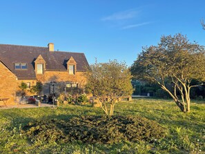 Ein Steinhaus mit einem geneigten Dach, einer kleinen Veranda und einem Garten mit verschiedenen Pflanzen und einem Baum.