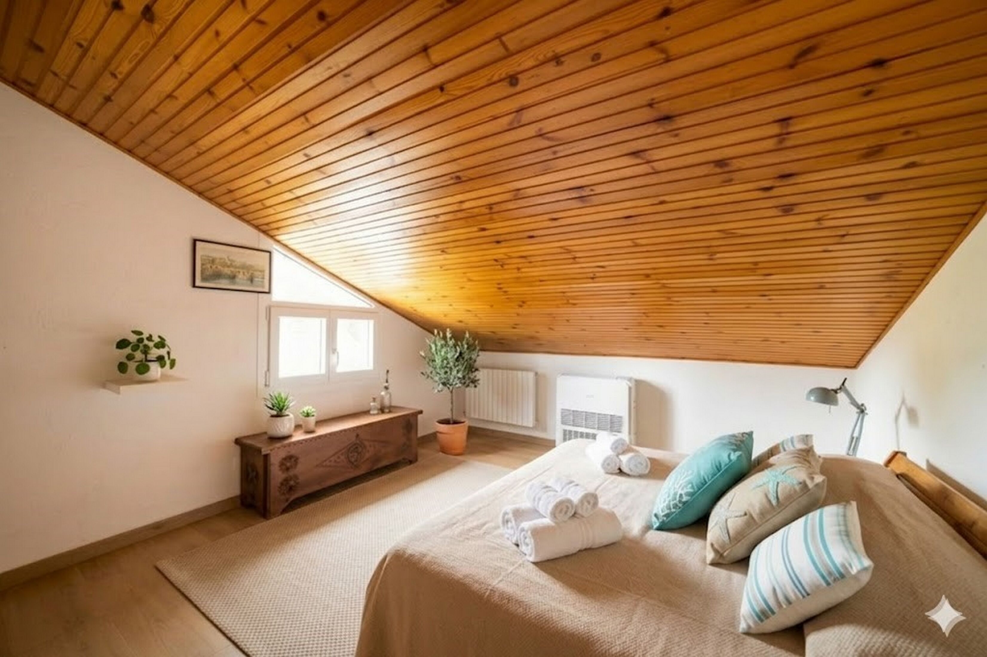 A bedroom with a sloped wooden ceiling, a bed with rolled towels, a dresser, a potted plant, and a window.