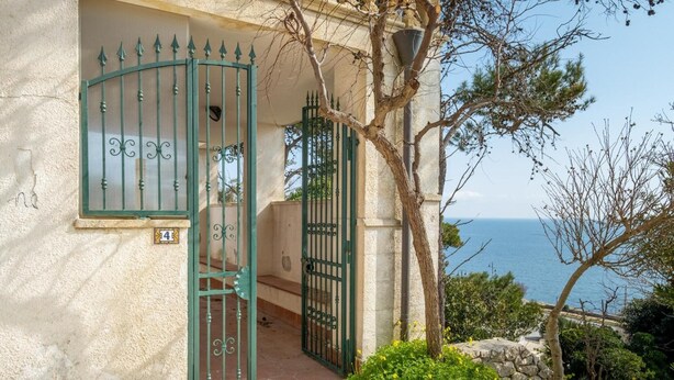 Un edificio costero con un portón verde, un muro de piedra y vista al mar.