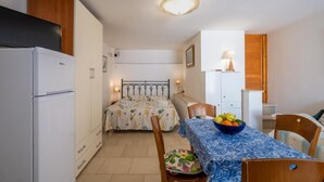 Habitación pequeña con cama, una mesita con frutero, refrigerador blanco y una lámpara.