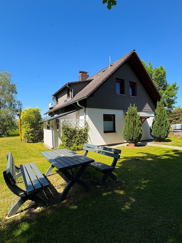 A two-story house with a sloped roof, a chimney, and a small attached garage. There is a picnic table in the front yard.