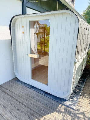 Een sauna met een glazen deur, een houten terras en een witte gevel.