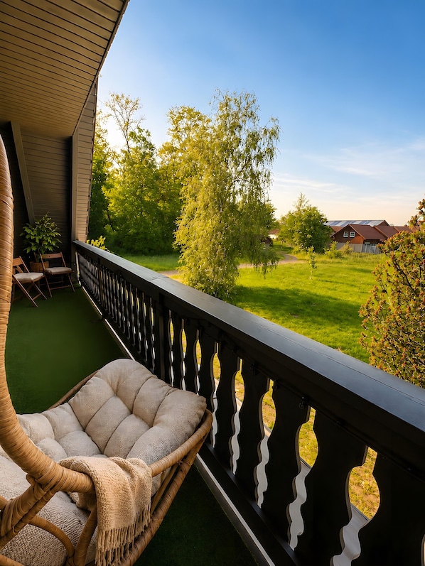 Ein Balkon mit einem Korbstuhl, einem kleinen Tisch und Blick auf eine grüne Wiese und Bäume.