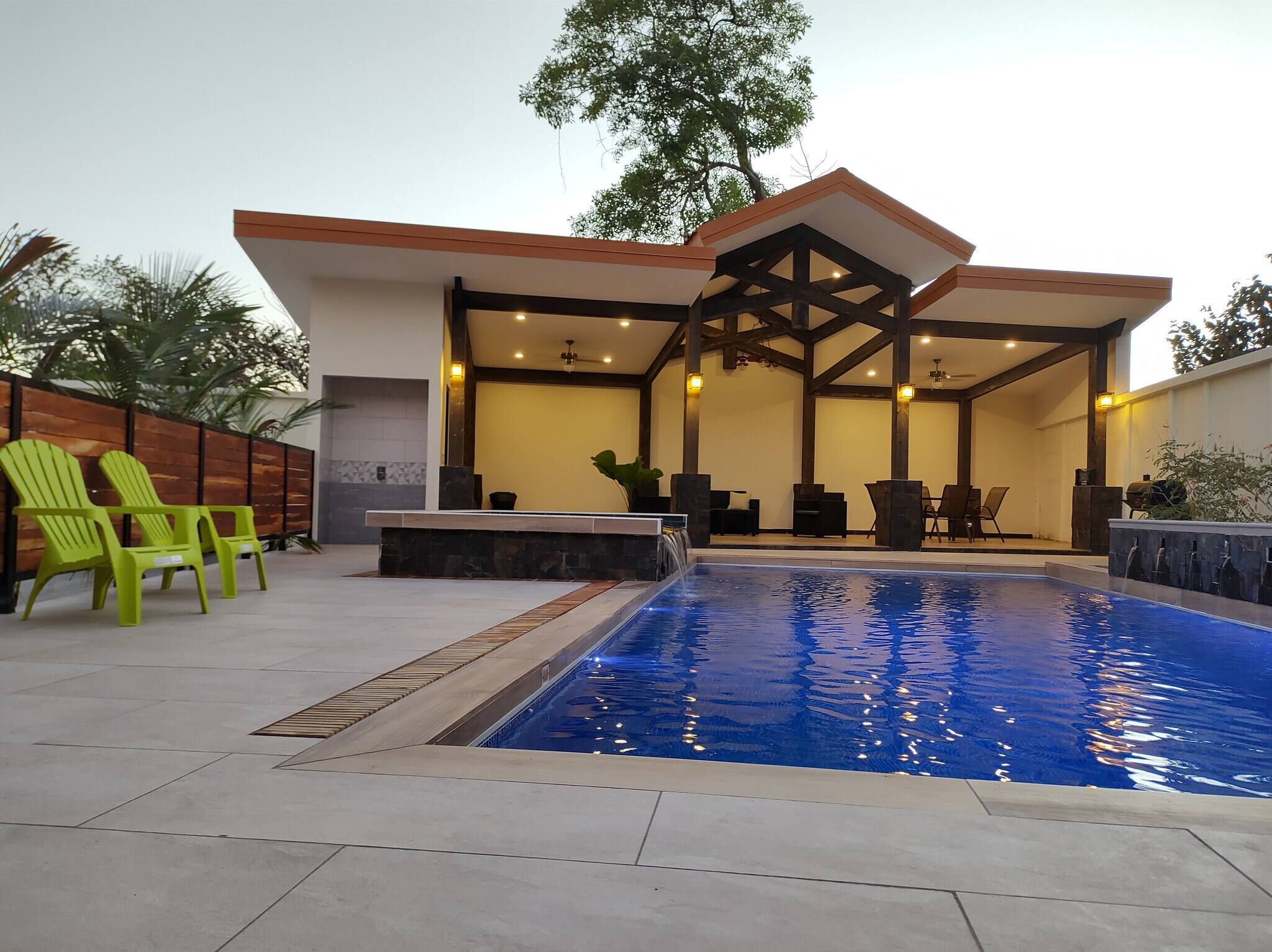 A modern outdoor pool area with a covered seating area, green chairs, and a stone wall.