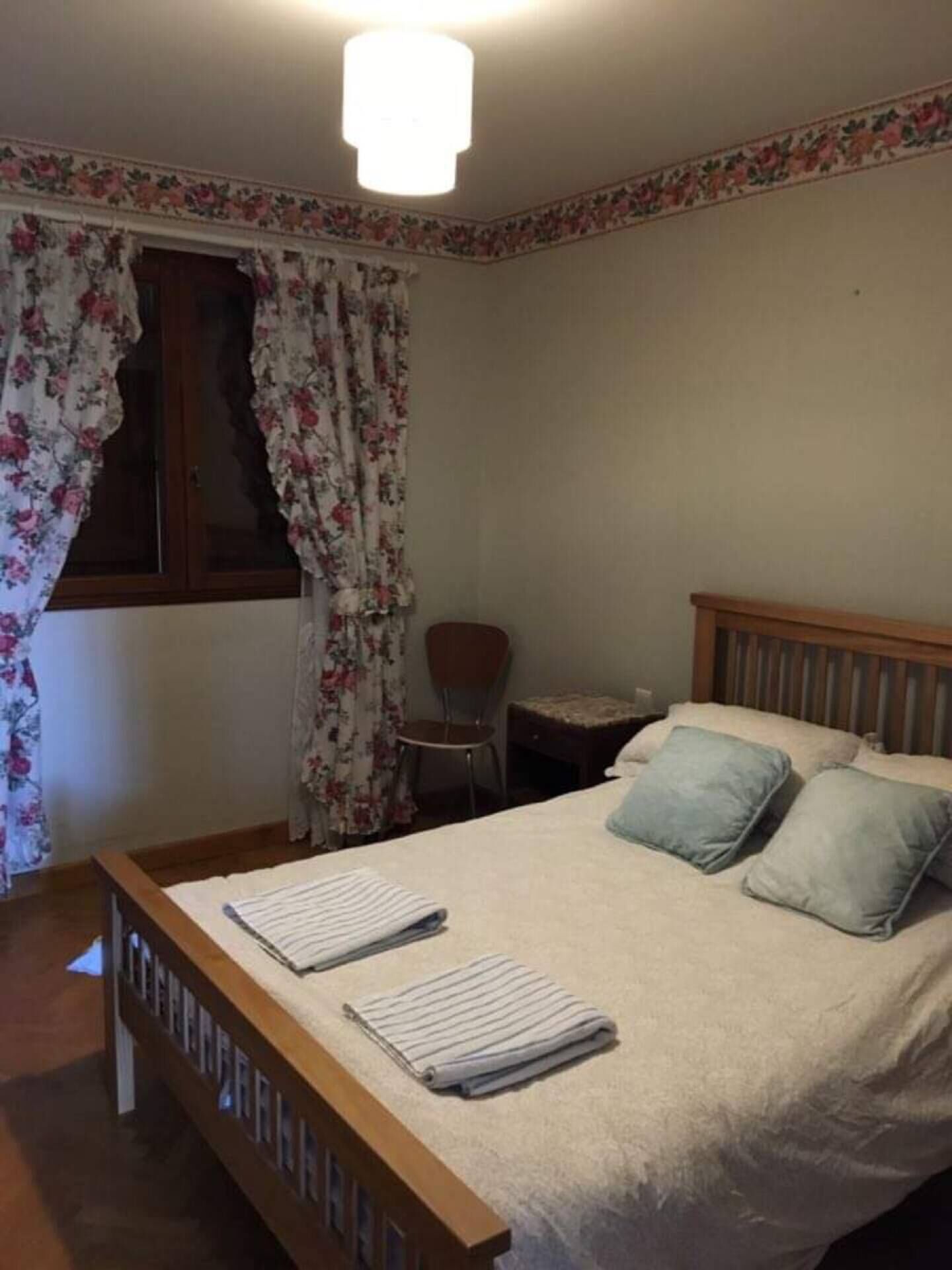 Een slaapkamer met een bed, kussens, een stoel, een klein tafeltje en een raam met bloemen gordijnen.