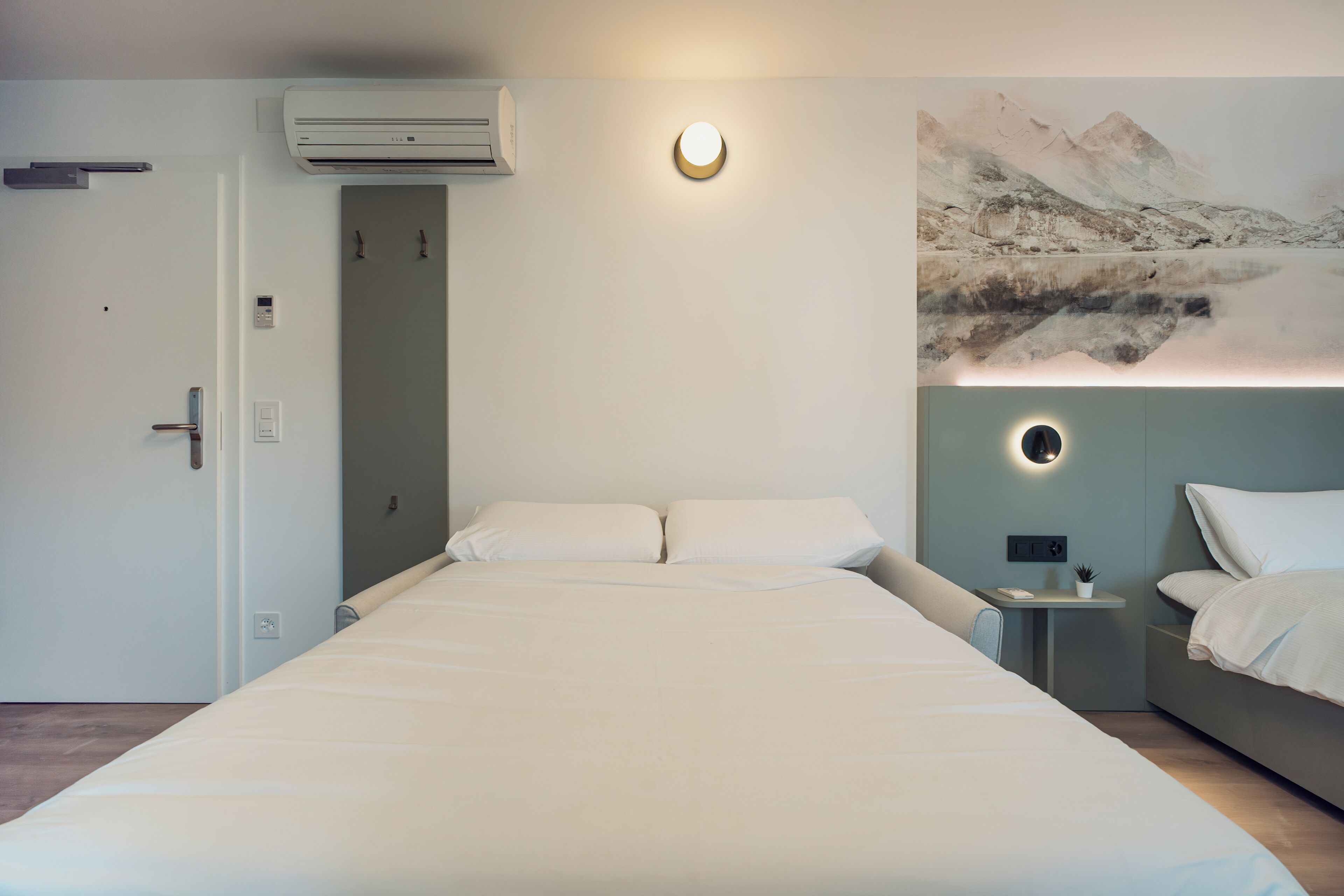 A modern hotel room with a large bed, a small bedside table, a wall-mounted air conditioner, and a painting of a mountain landscape.