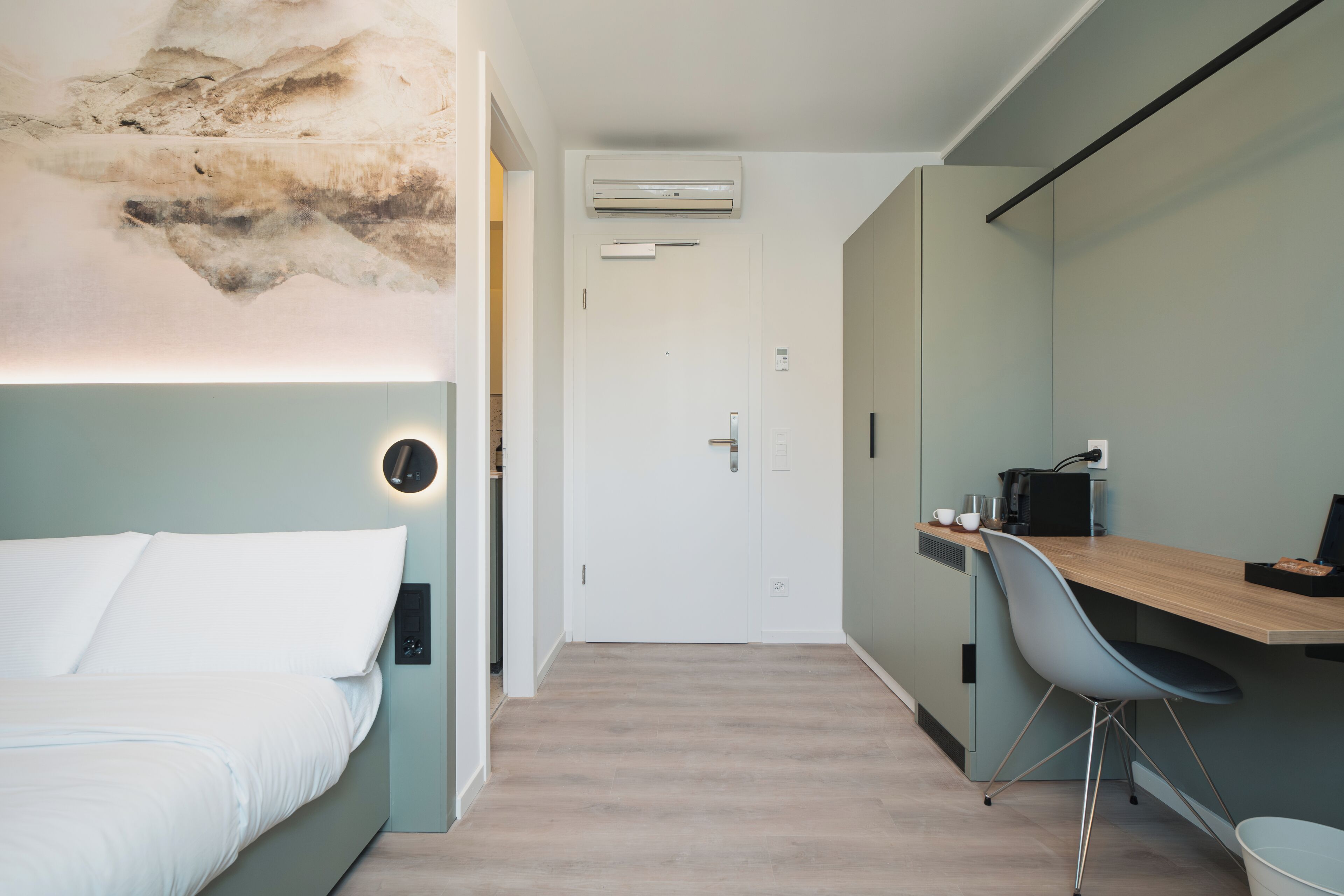 A modern hotel room with a bed, a desk, a chair, and an air conditioning unit.