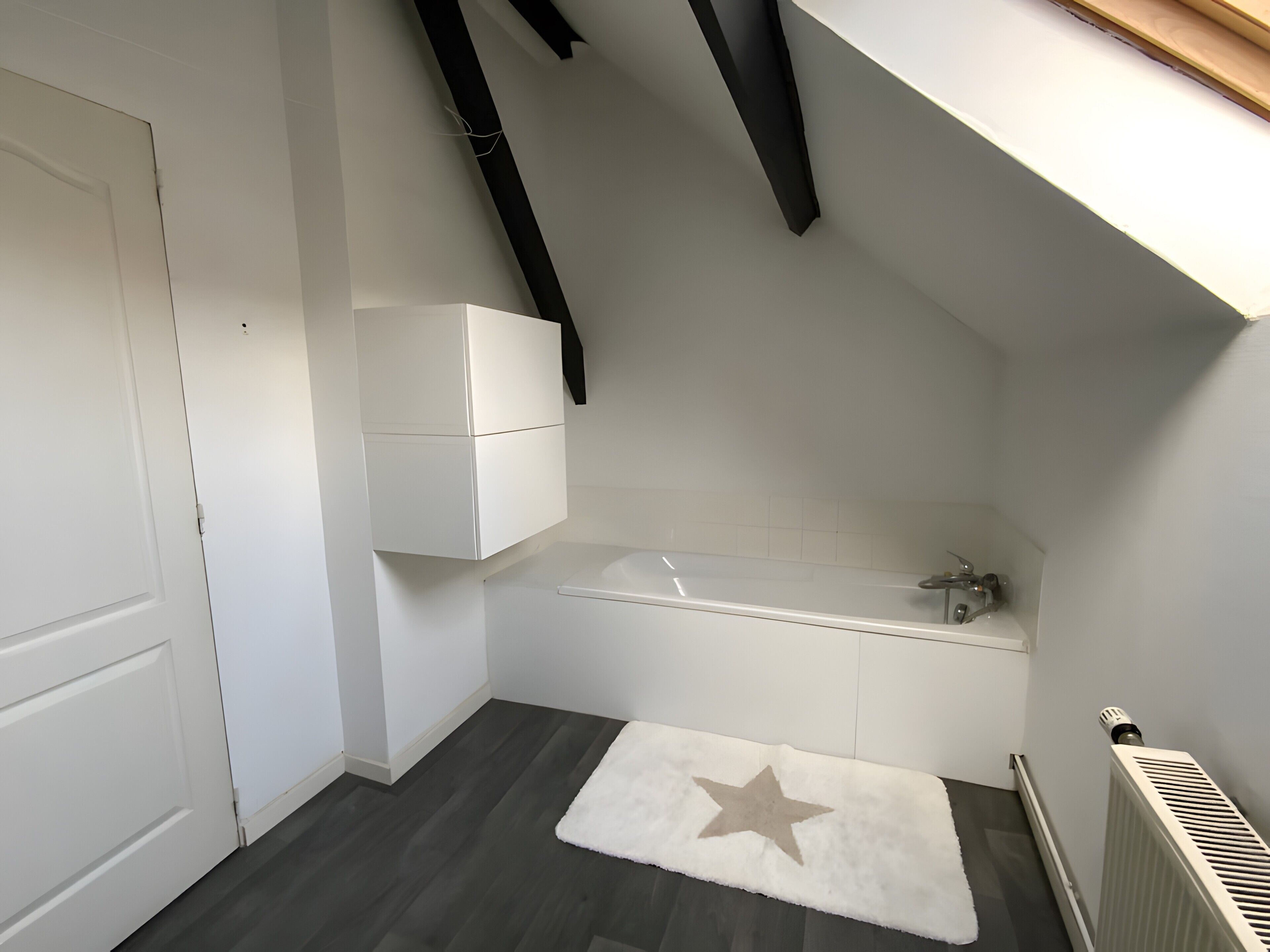 Een kamer met een witte deur, een witte kast, een wit bad en een vloerkleed met stervenspatroon, en een radiator.