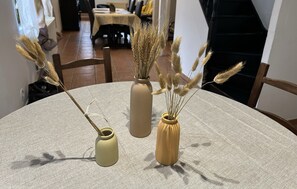 Ein runder Tisch mit drei Vasen, gefüllt mit getrockneten Pflanzen, ein Holzstuhl und im Hintergrund ein Essbereich.