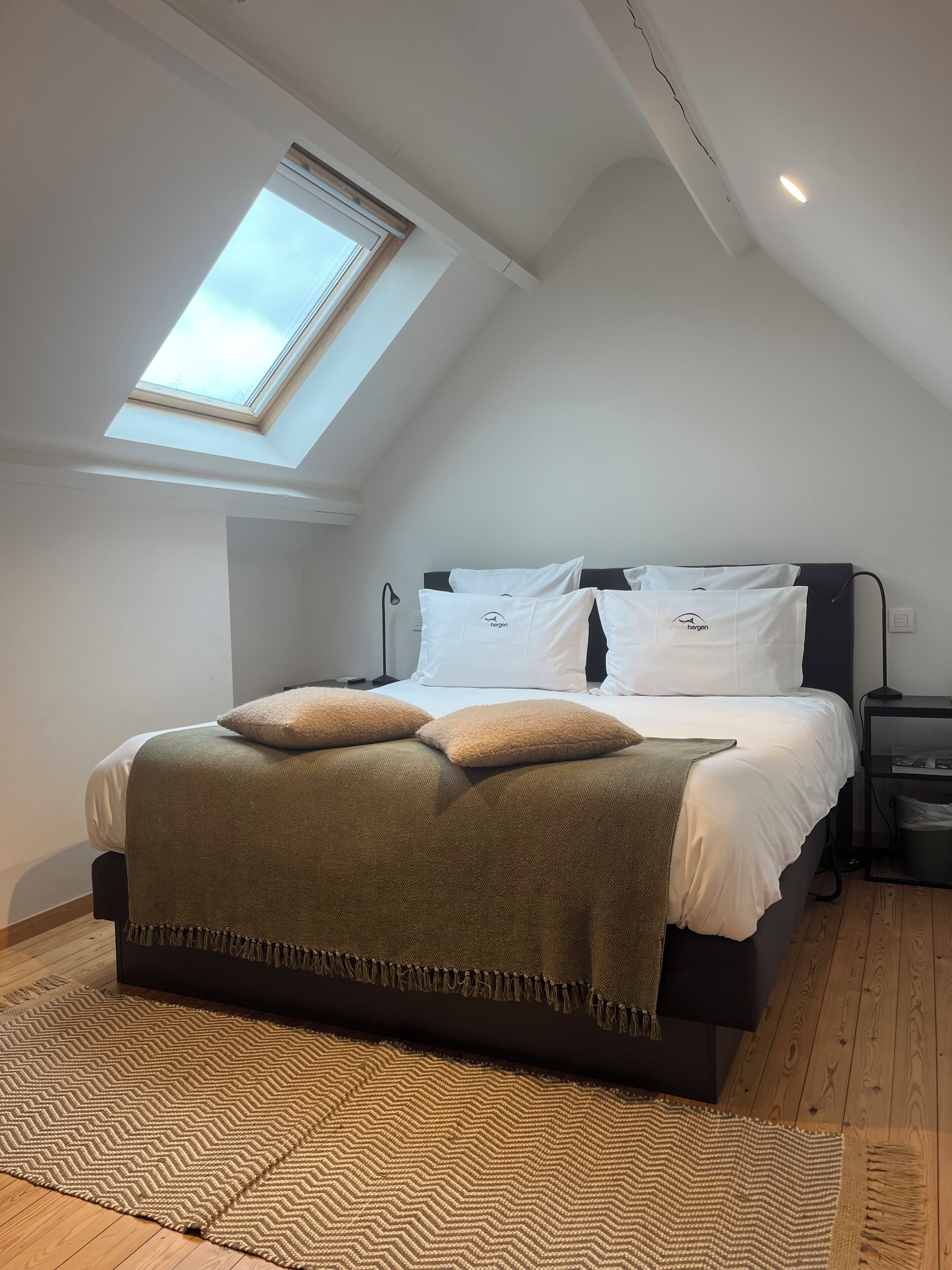Ein Schlafzimmer mit einem Dachfenster, einem Bett mit weißen Leinen und zwei Kissen, einem dunklen Kopfteil und einem kleinen Nachttisch mit einer Lampe.
