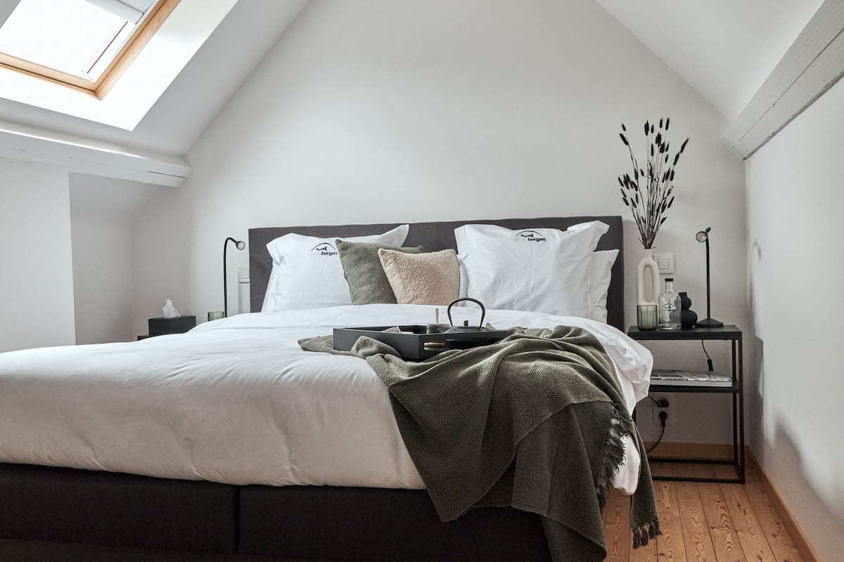 A bedroom with a skylight, a bed with a dark headboard, a bedside table with a lamp, and a wooden floor.