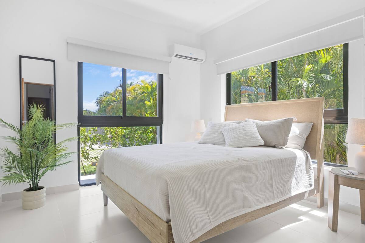 A modern bedroom with a large bed, a potted plant, and a view of greenery through large windows.