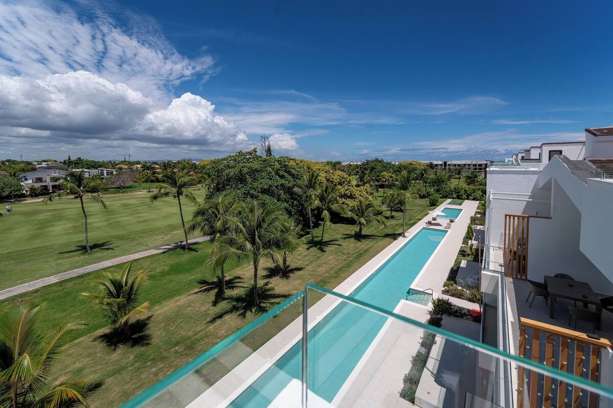 A modern hotel with a swimming pool, lush greenery, and a clear blue sky.