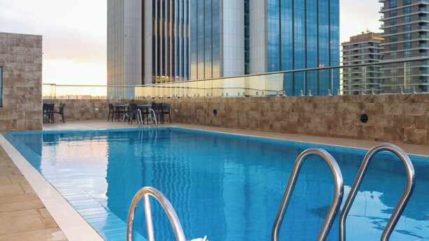 A swimming pool with metal railings, a stone wall, and a view of tall buildings in the background. - Elite Residence 1 BR: Balcony, Heated Pool, Power backup, Sky Gym& 24/7 Security (Nairobi)