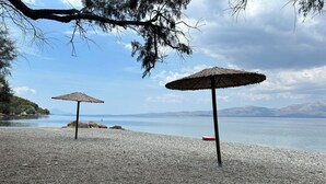 Ein Kiesstrand mit zwei reetgedeckten Sonnenschirmen und einem roten Schwimmreifen.