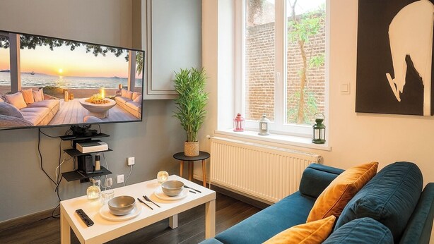 Un salon avec un téléviseur diffusant une vue sur un coucher de soleil, un canapé, une petite table avec un bol et des baguettes, et une plante en pot près de la fenêtre.