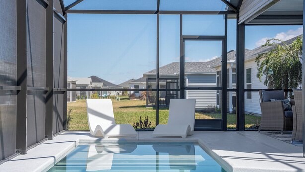Uma área de piscina moderna ao ar livre, com paredes de vidro transparente, duas espreguiçadeiras brancas e vista para casas vizinhas.