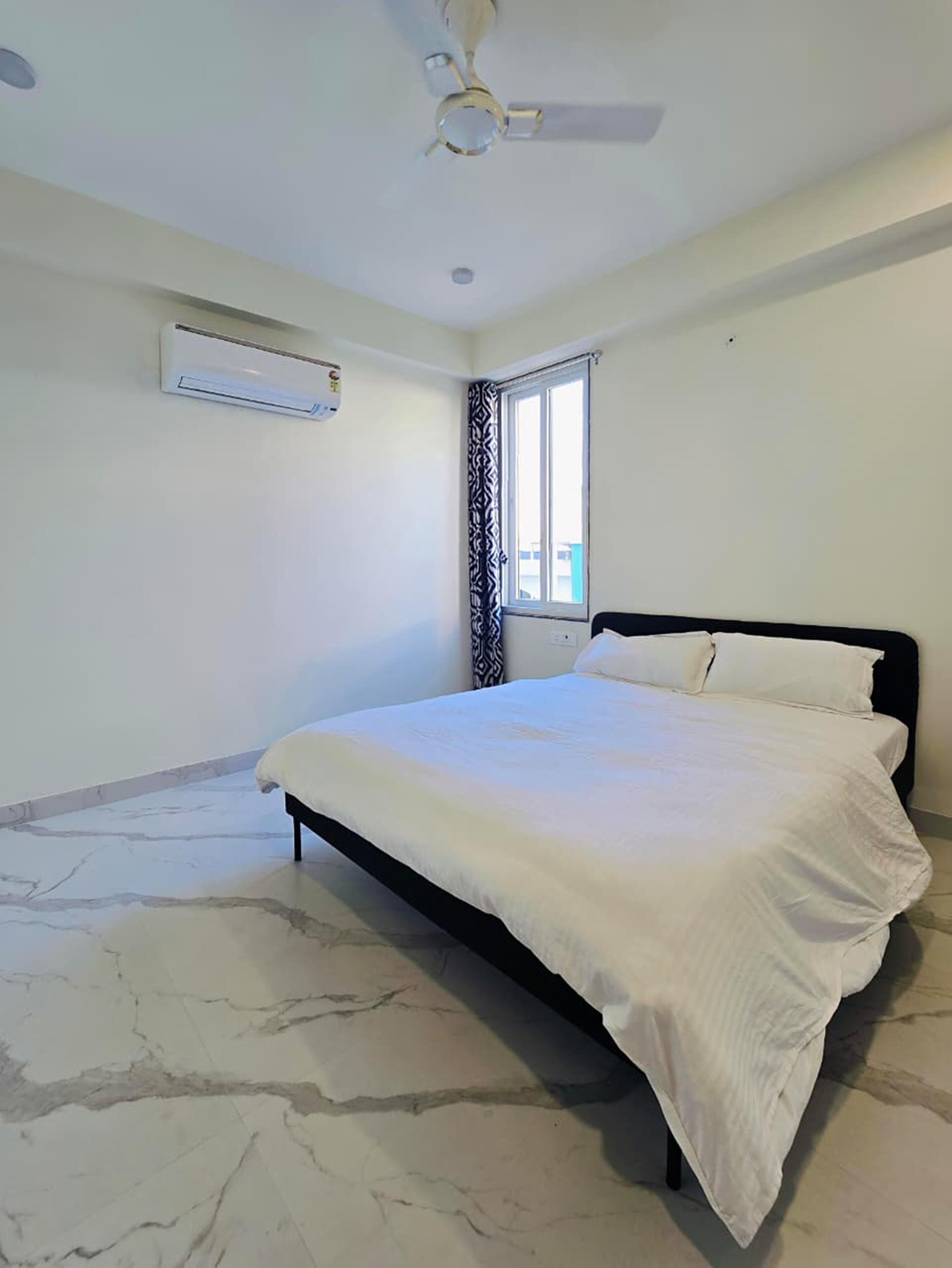 Un dormitorio con cama, ventilador de techo, aire acondicionado, ventana con cortina estampada y piso con diseño de mármol.