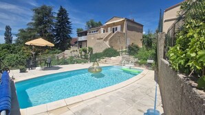 Una piscina con tobogán, rodeada de una terraza de piedra y una valla, con una casa al fondo.