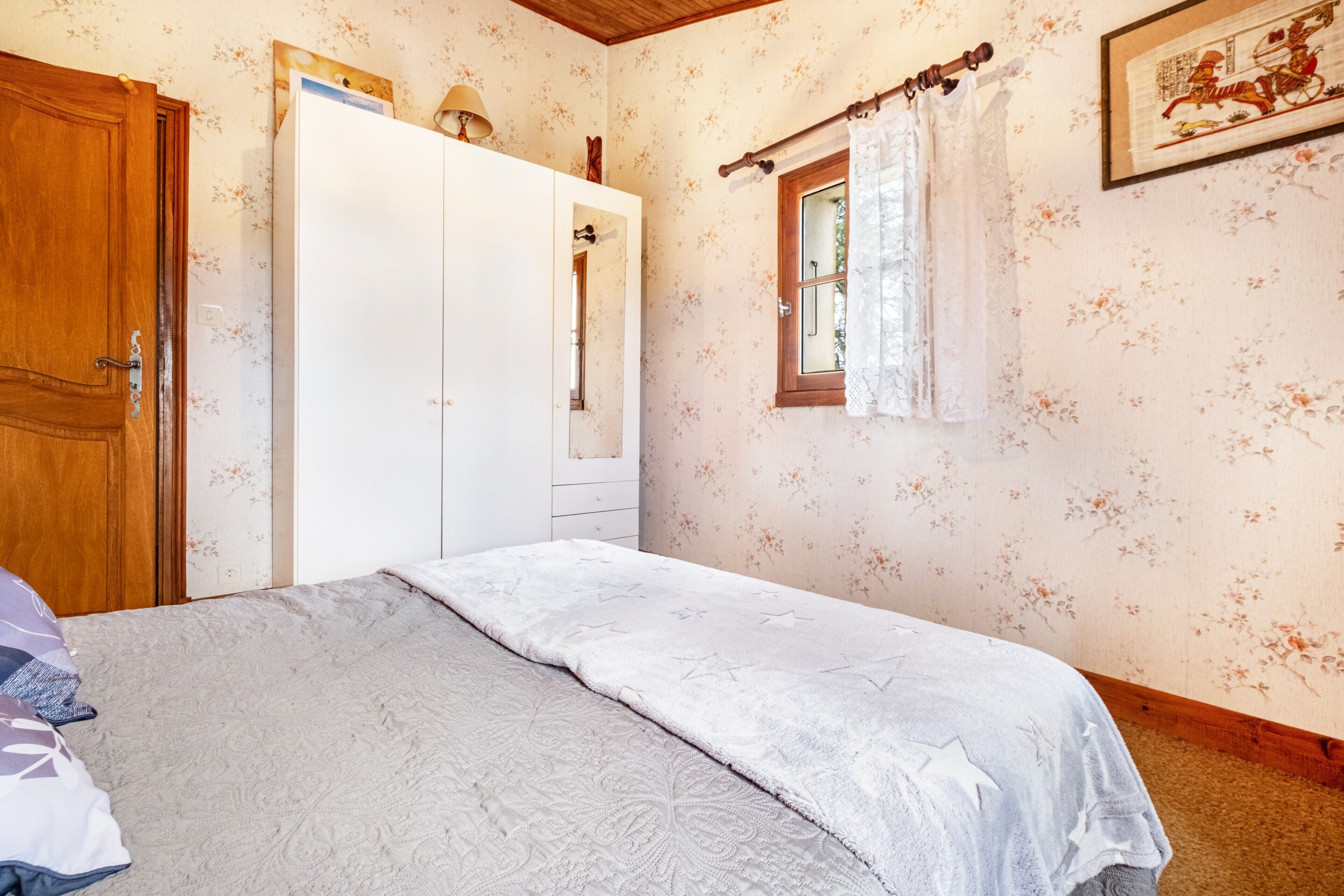 Una camera da letto con un letto, un armadio, una finestra con tende e una parete con carta da parati a motivi floreali.