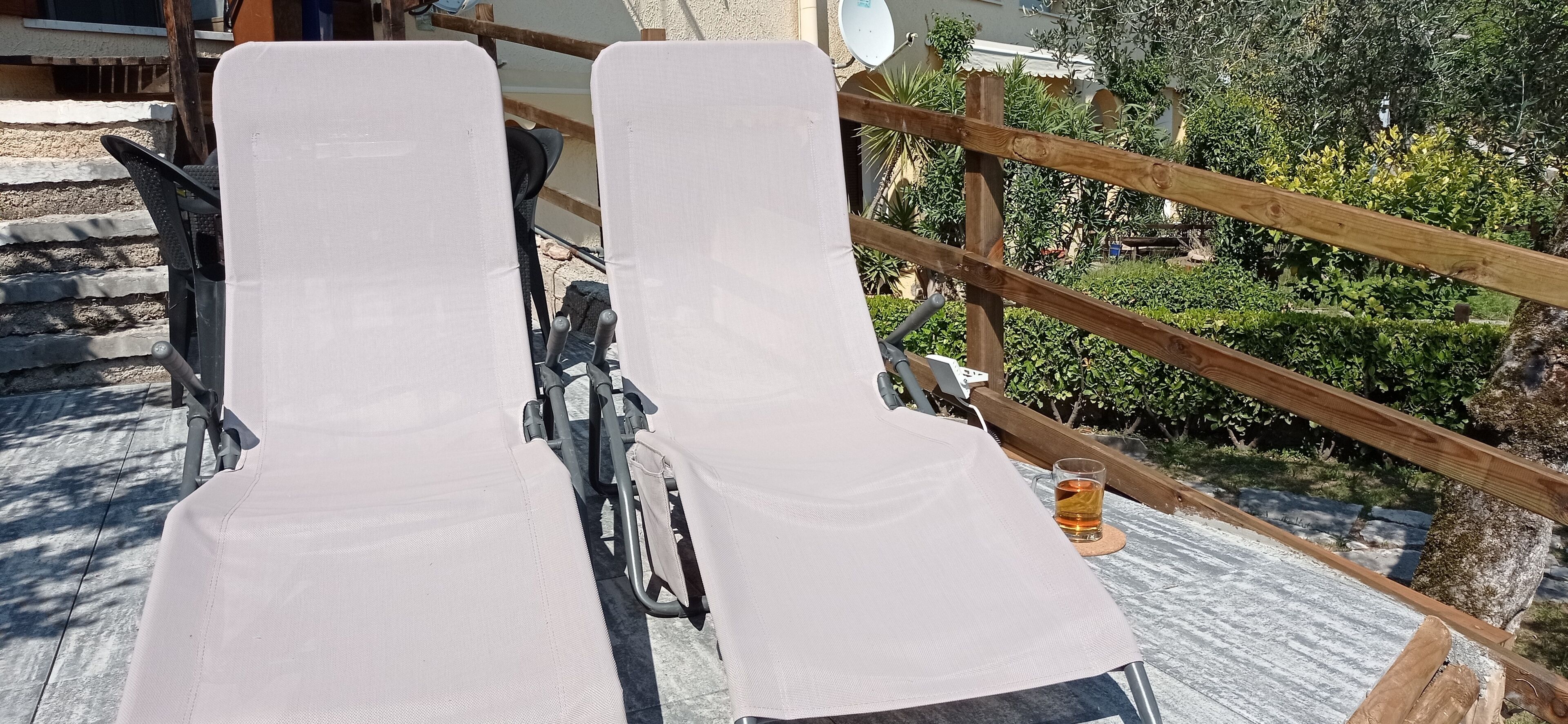 Deux fauteuils de jardin blancs à structure métallique, une rambarde en bois et une table en verre avec une boisson.