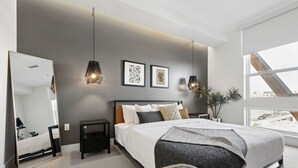 A modern bedroom with a large bed, a bedside table, a nightstand, a mirror, and a window with a view of the street.