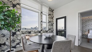 A modern dining area with a round table, grey upholstered chairs, a potted plant, and a city view through large windows. - Top Floor | 1BR Corner Loft | LoHi Denver | Dog-Friendly (Denver)