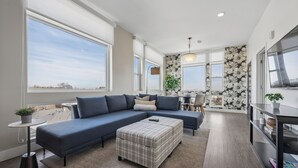 A modern living room with a large sectional sofa, a small round table with a plant, and a view of the city through large windows. - Top Floor | 1BR Corner Loft | LoHi Denver | Dog-Friendly (Denver)