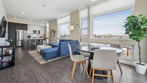 A modern kitchen with stainless steel appliances, a dining area with a round table and chairs, a blue sofa, and large windows offering a city view. - Top Floor | 1BR Corner Loft | LoHi Denver | Dog-Friendly (Denver)
