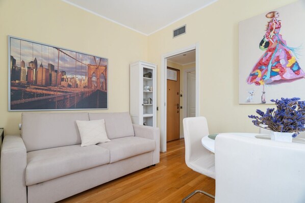 Una sala de estar con un sofá, un cuadro de un paisaje urbano, un aparador blanco y un comedor con una mesa y sillas.